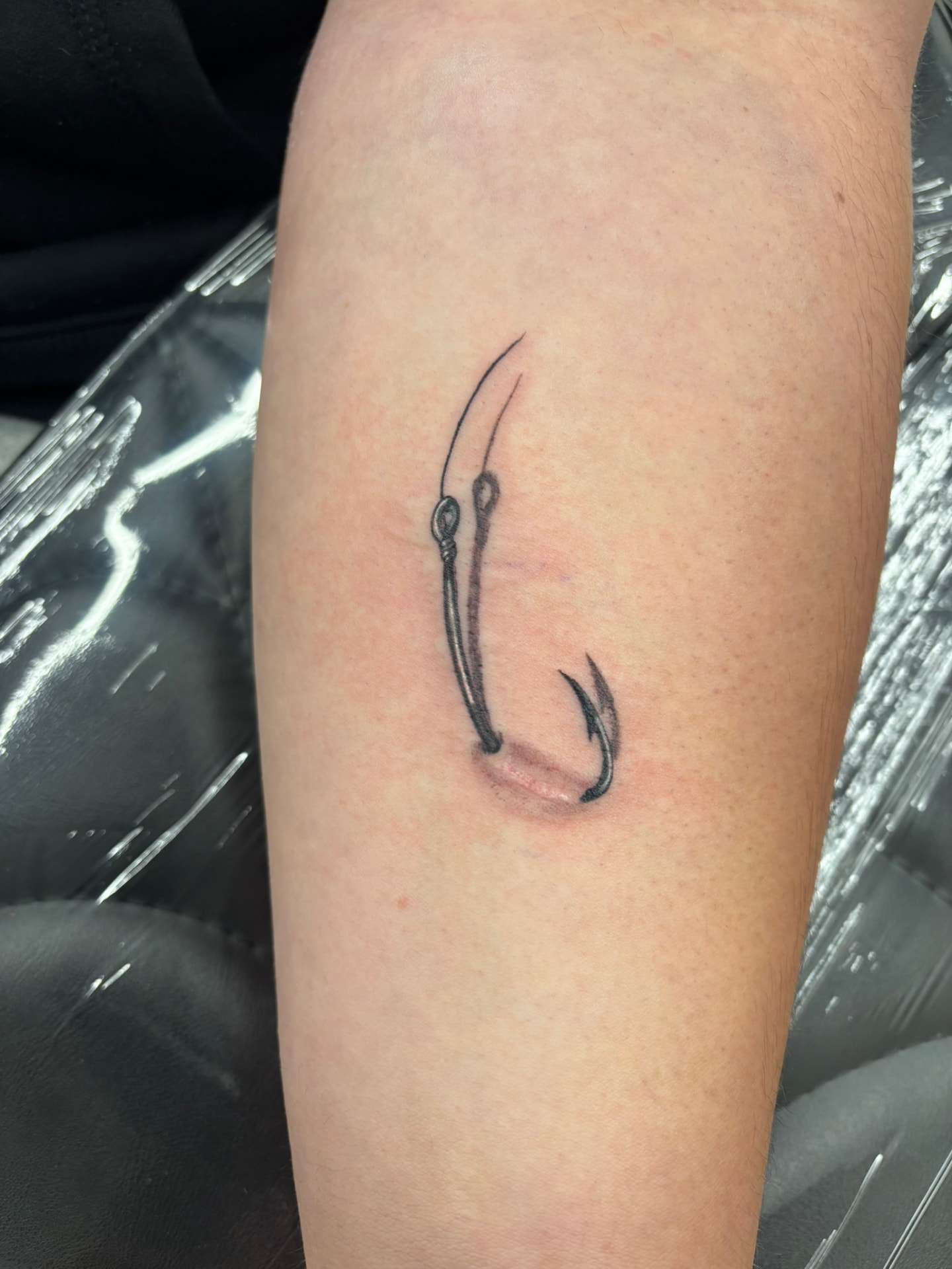 Black and grey tattoo of two fishing hooks on lines, one shaded to look as if it hooks through the skin