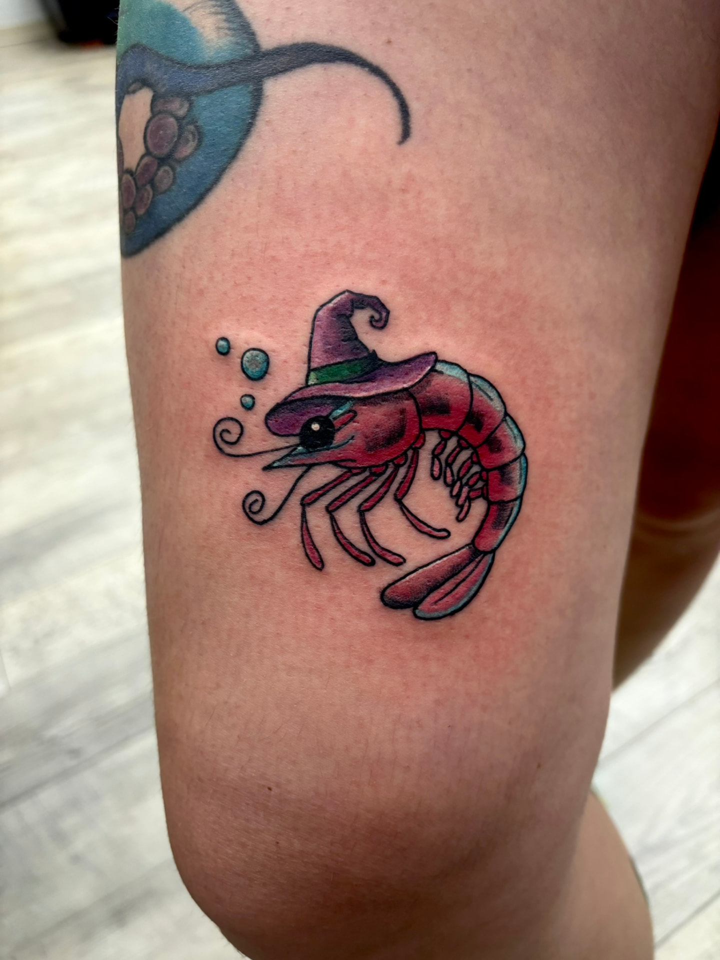 Color tattoo of a shrimp wearing a purple witch hat with floating bubbles on the lower thigh