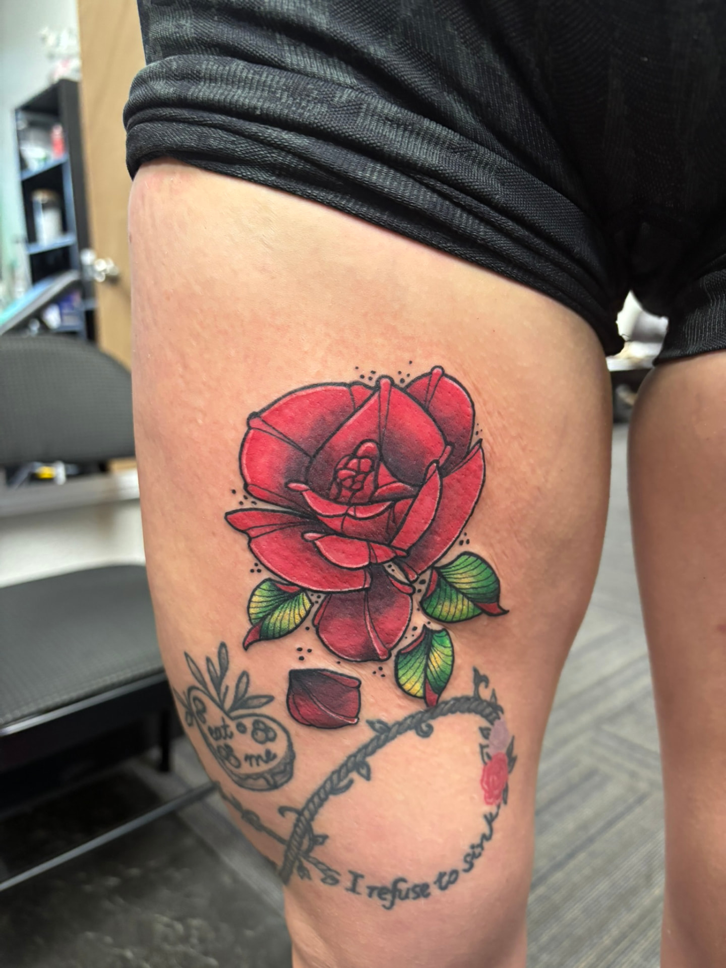 Color tattoo of a red rose in full bloom with green leaves and a single fallen petal on the thigh