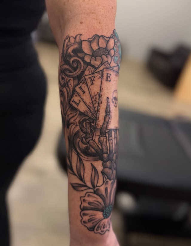 Black and grey forearm tattoo of a skeleton hand holding cards spelling LIFE, with flowers and filigree
