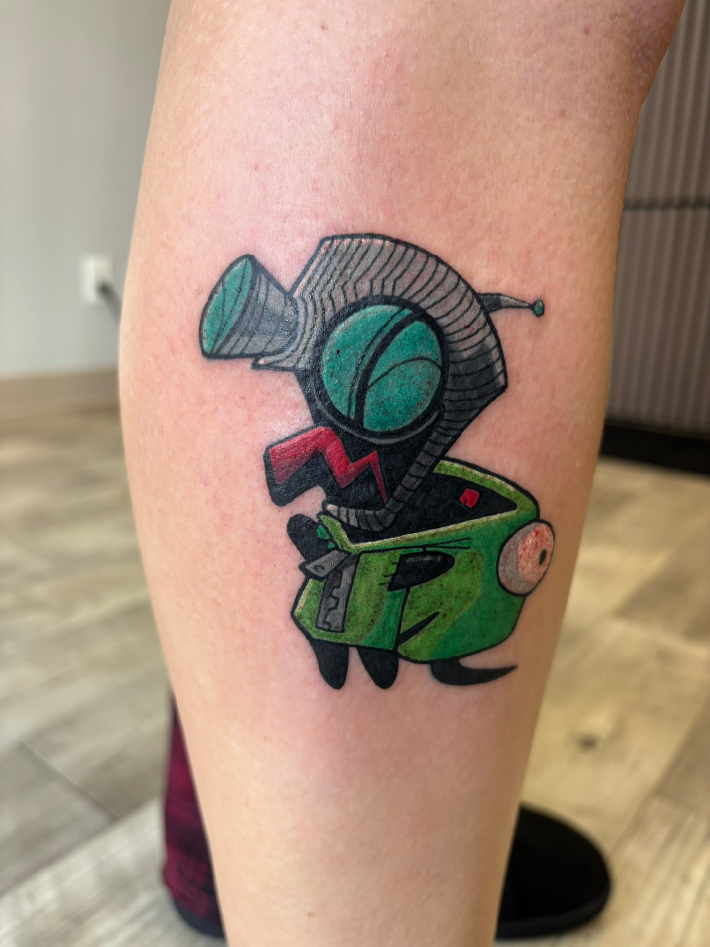 Color cartoon tattoo of a robot unzipping a green dog suit costume to reveal its metal head and antenna on the calf