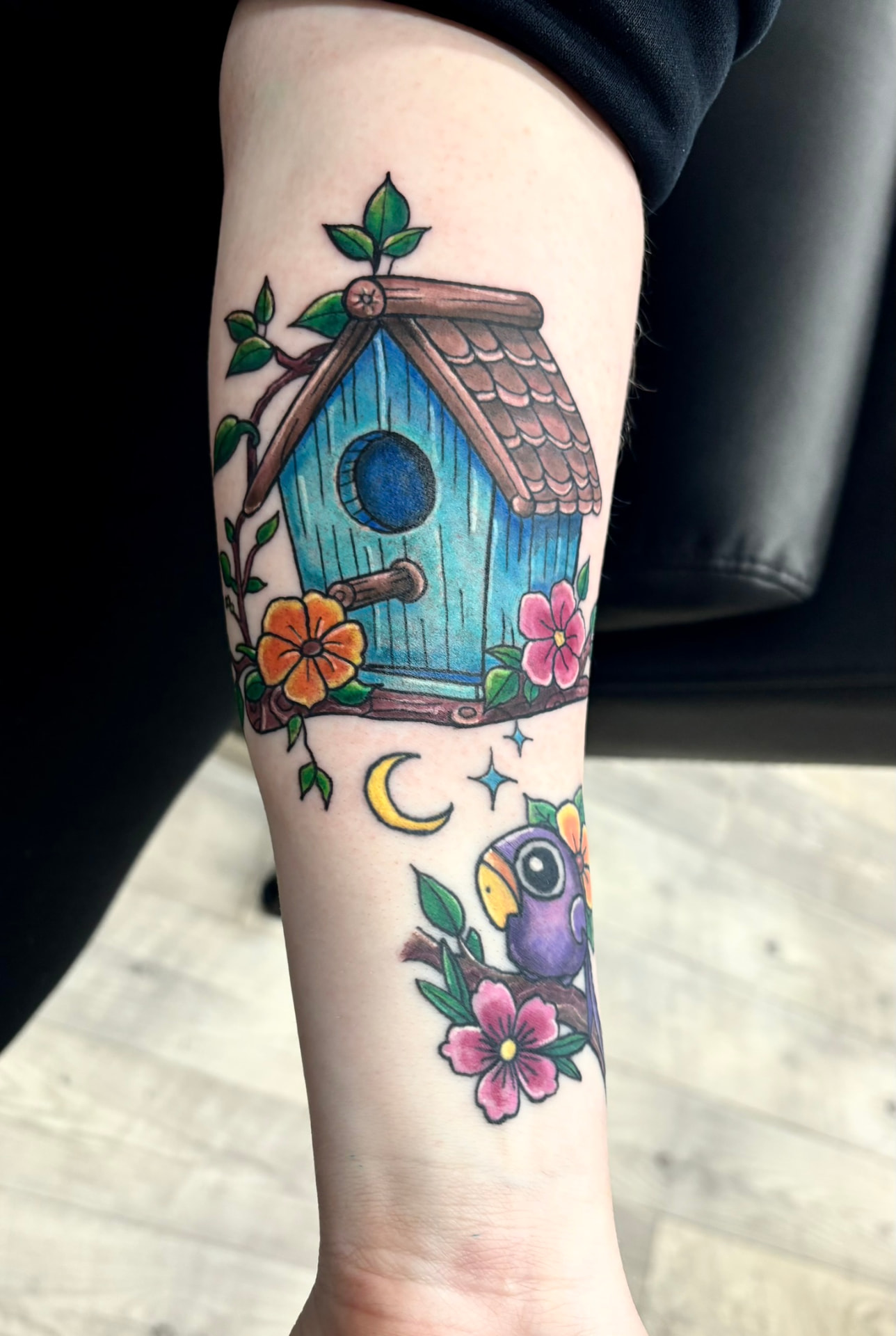 Color tattoo of a blue birdhouse on flowering branches framed by a crescent moon, stars, and a small purple bird on a forearm