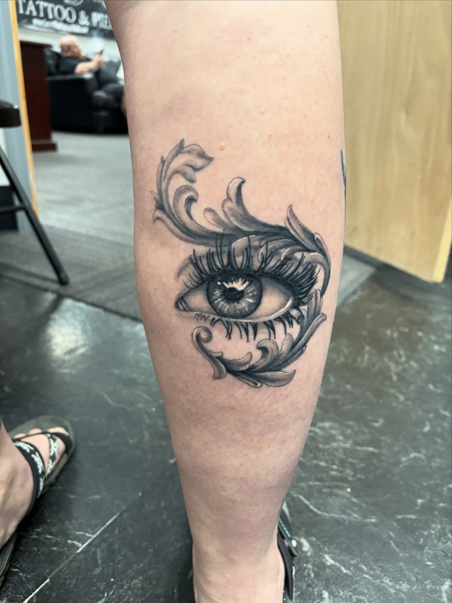 Black and grey tattoo of a realistic human eye framed by ornate baroque filigree leaves on the lower leg