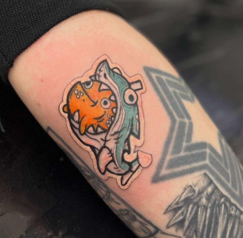 Cartoon sticker-style tattoo of an orange creature wearing a teal shark costume
