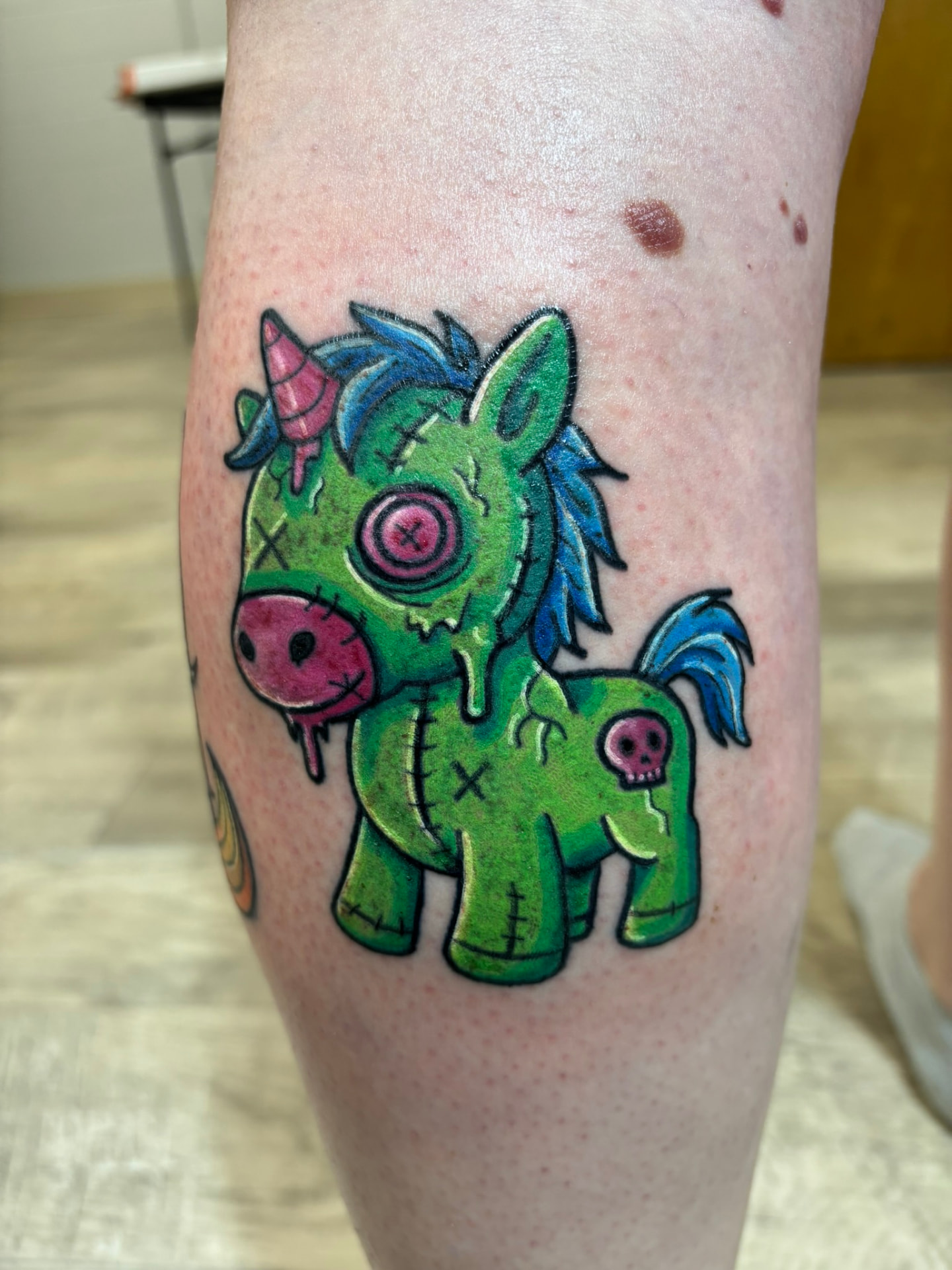 Color cartoon tattoo of a stitched green plush unicorn with a pink button eye and dripping slime on the calf