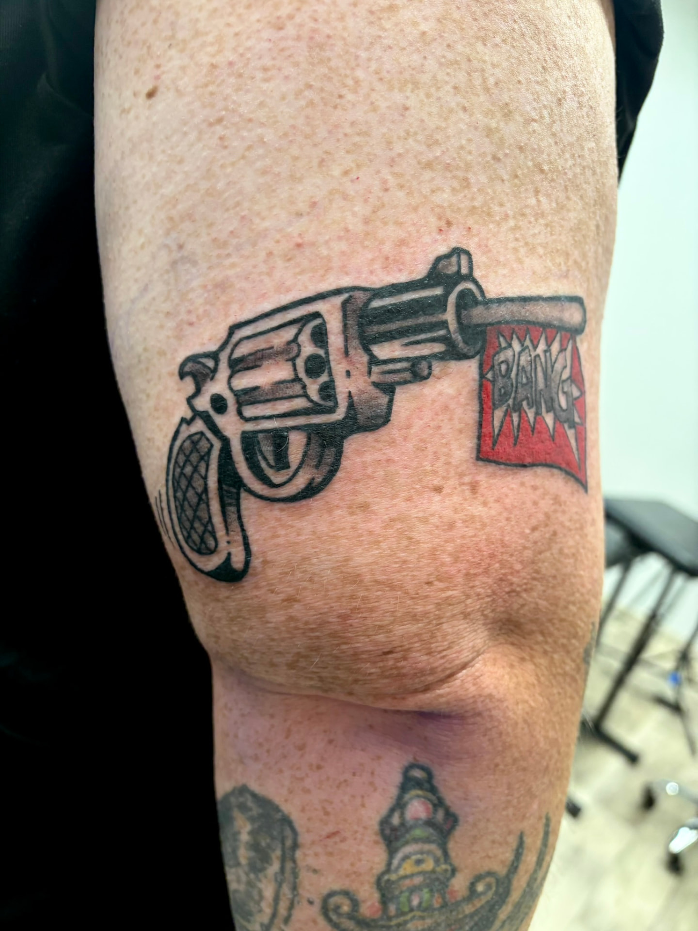 Traditional tattoo of a revolver firing a red flag reading BANG from a burst bubble on the upper arm