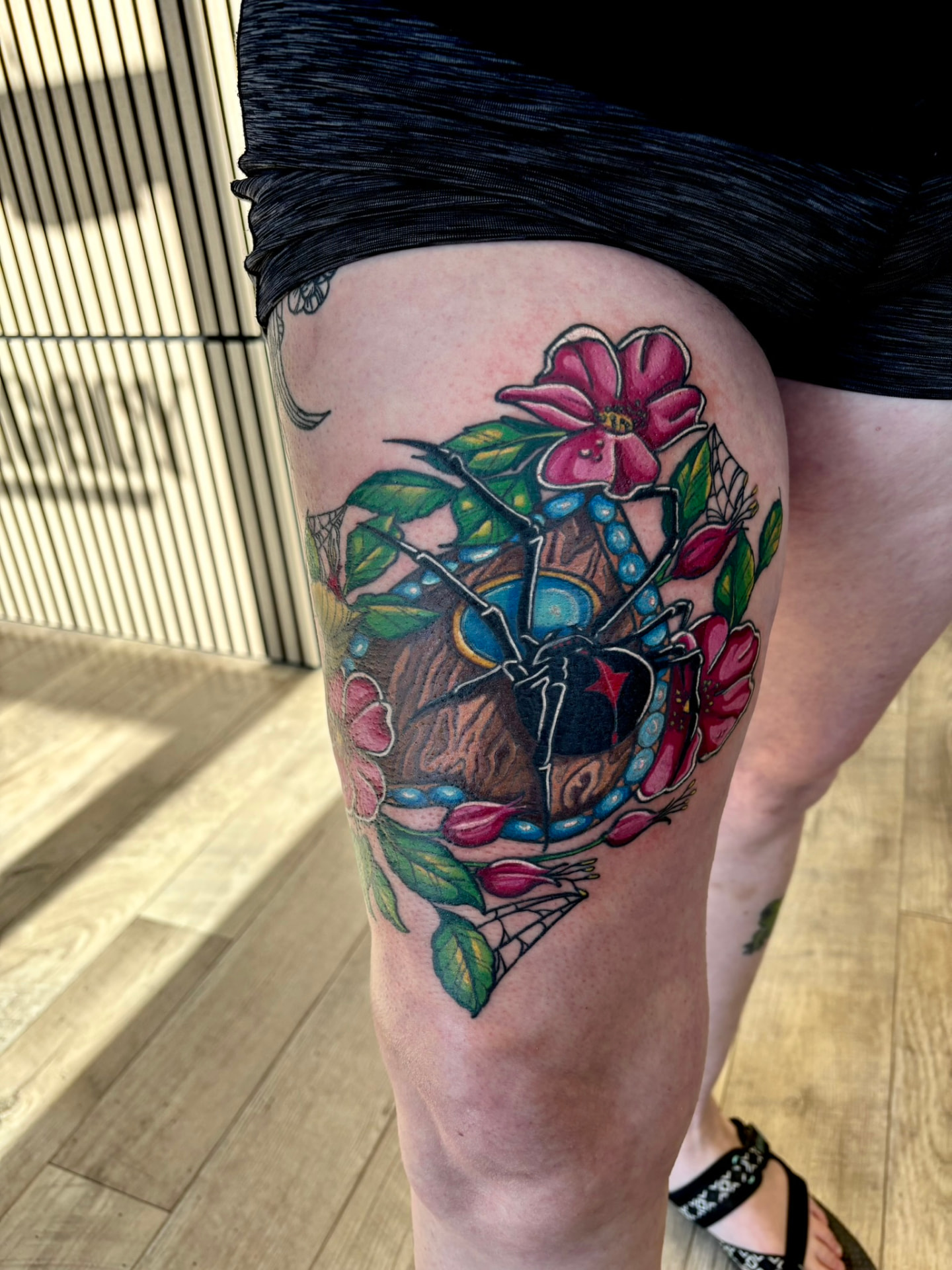 Neo-traditional color tattoo of a black widow spider inside a beaded oval frame surrounded by pink roses and cobwebs